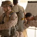 Two military personnel in desert camouflage uniforms are engaged in a parachute equipment inspection or fitting procedure. One soldier wearing a tactical helmet and parachute pack stands still while a second soldier closely inspects the parachute harness and pack from behind. Additional personnel and a whiteboard with writing are visible in the background.