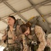 Two military personnel in desert camouflage uniforms (MARPAT pattern consistent with U.S. Marines) are wearing parachute harnesses and parachute packs, appearing to prepare for a airborne jump operation. The female service member in the foreground is looking down and adjusting or checking a buckle or fastener on her harness, while the male service member behind her looks upward. They are standing inside what appears to be a hangar or staging area.