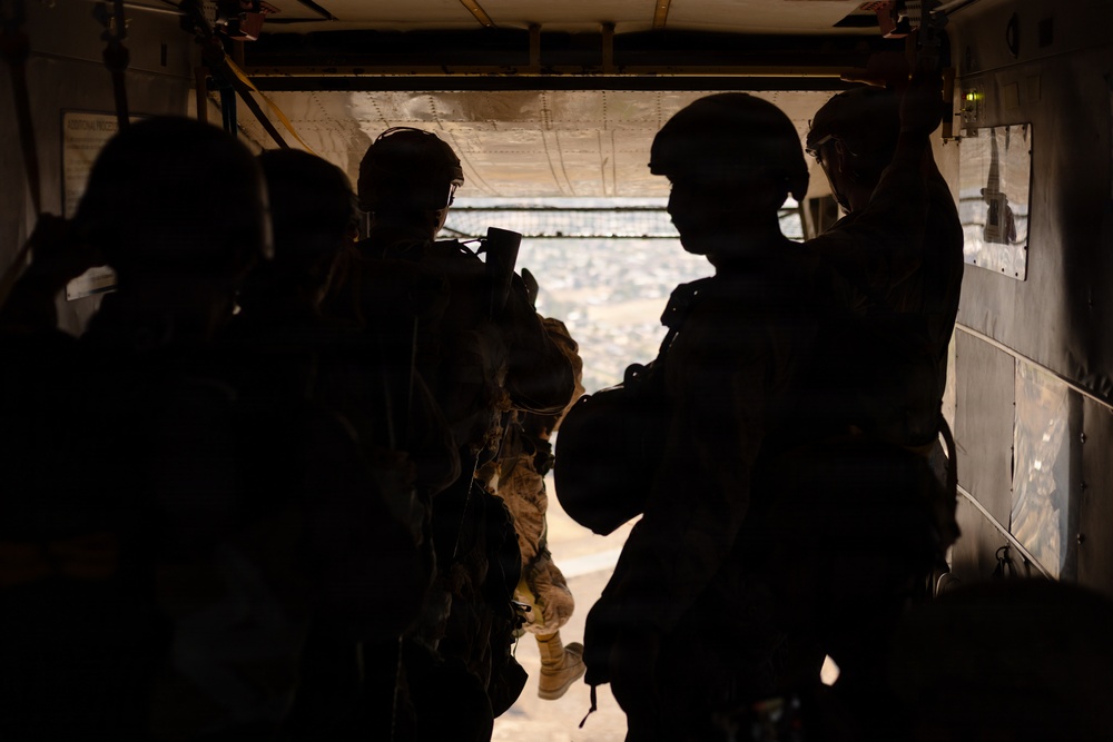 The image shows silhouetted military personnel standing at the open rear ramp of what appears to be a military transport helicopter, likely a CH-47 Chinook, at altitude. The figures are wearing combat helmets and tactical gear, positioned near the open ramp with a landscape visible below. The strong backlight creates a high-contrast silhouette effect, obscuring individual details.