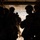 The image shows silhouetted military personnel standing at the open rear ramp of what appears to be a military transport helicopter, likely a CH-47 Chinook, at altitude. The figures are wearing combat helmets and tactical gear, positioned near the open ramp with a landscape visible below. The strong backlight creates a high-contrast silhouette effect, obscuring individual details.