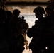 The image shows silhouetted military personnel standing at the open rear ramp of what appears to be a military transport helicopter, likely a CH-47 Chinook, at altitude. The figures are wearing combat helmets and tactical gear, positioned near the open ramp with a landscape visible below. The strong backlight creates a high-contrast silhouette effect, obscuring individual details.