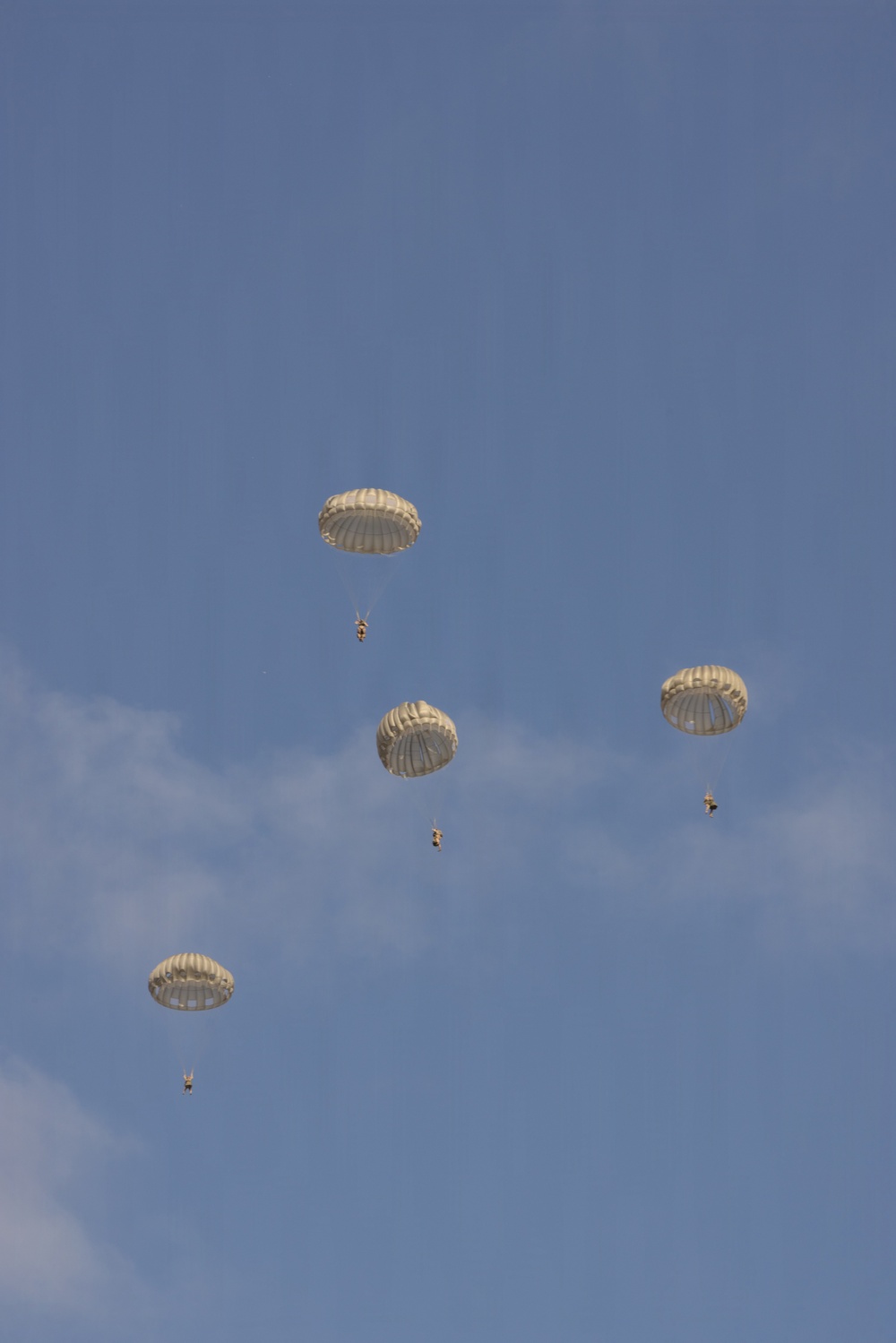 The image shows multiple military-style paratroopers descending through a partly cloudy blue sky. Three fully deployed round parachutes are clearly visible, each with a suspended figure beneath them. The parachutes appear to be standard military round canopy design in a tan or off-white color.