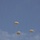 The image shows multiple military-style paratroopers descending through a partly cloudy blue sky. Three fully deployed round parachutes are clearly visible, each with a suspended figure beneath them. The parachutes appear to be standard military round canopy design in a tan or off-white color.