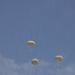 The image shows multiple military-style paratroopers descending through a partly cloudy blue sky. Three fully deployed round parachutes are clearly visible, each with a suspended figure beneath them. The parachutes appear to be standard military round canopy design in a tan or off-white color.