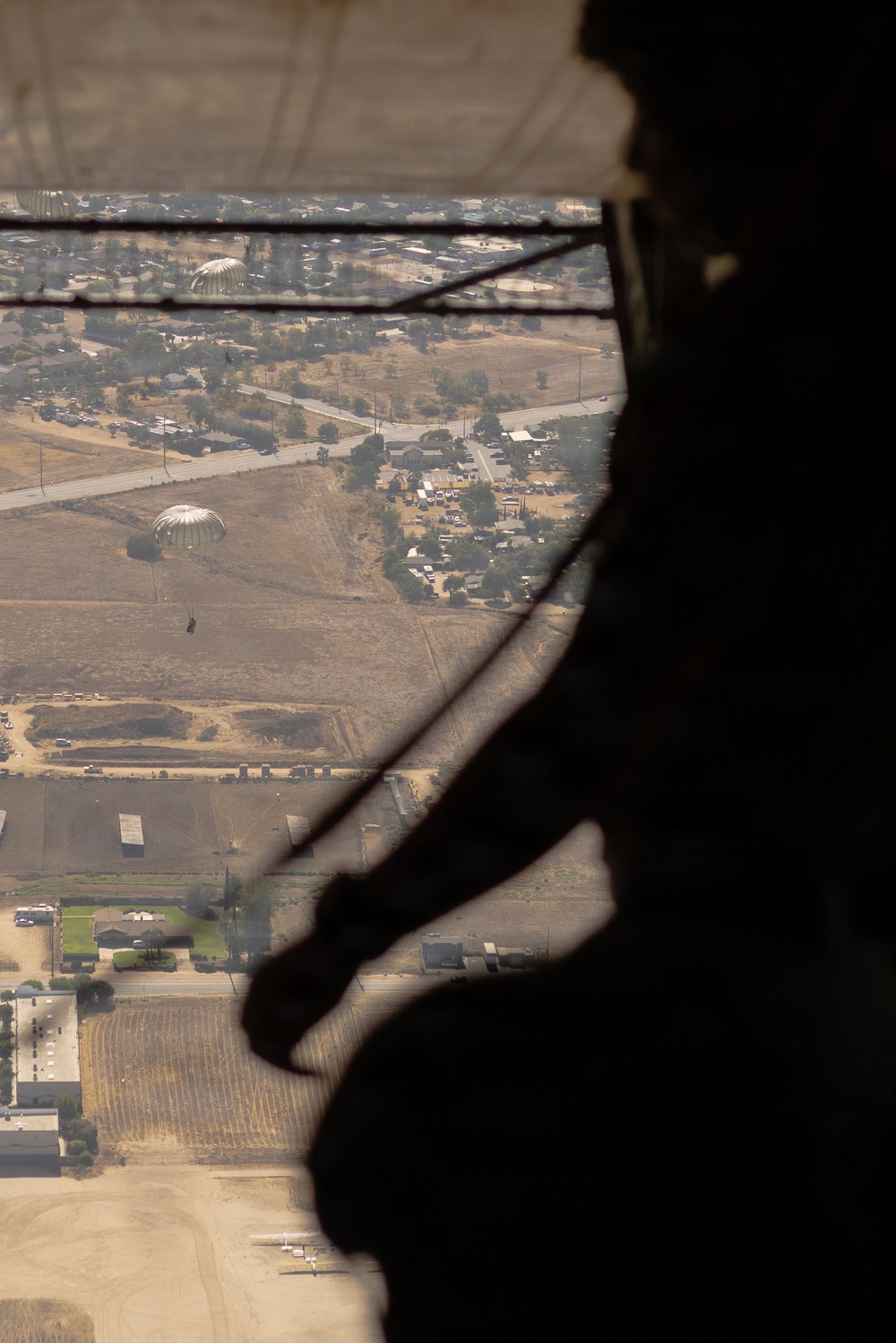 The image is taken from inside a military aircraft looking out through an open door or ramp at an aerial view of a semi-arid landscape below. Two deployed parachutes with suspended jumpers are visible descending toward the ground, indicating an active airborne/paratrooper operation. A silhouetted figure, likely a jumpmaster or crew member, is visible in the foreground right side of the frame.