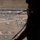The image is taken from inside a military aircraft looking out through an open door or ramp at an aerial view of a semi-arid landscape below. Two deployed parachutes with suspended jumpers are visible descending toward the ground, indicating an active airborne/paratrooper operation. A silhouetted figure, likely a jumpmaster or crew member, is visible in the foreground right side of the frame.