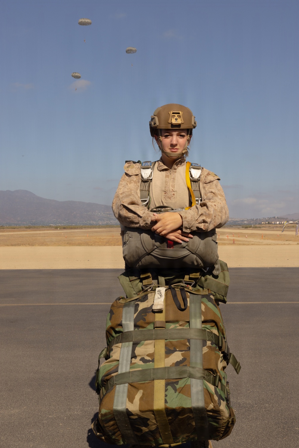 A military service member in full parachute jump gear stands on a tarmac or airfield, posing with arms crossed in front of a large camouflage kit bag. Multiple parachutes are visible descending in the background sky, indicating an active airborne operation. The individual is wearing USMC-style MARPAT desert utilities, a tactical helmet with NVG mount, and a parachute harness with reserve.