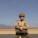 A military service member in full parachute jump gear stands on a tarmac or airfield, posing with arms crossed in front of a large camouflage kit bag. Multiple parachutes are visible descending in the background sky, indicating an active airborne operation. The individual is wearing USMC-style MARPAT desert utilities, a tactical helmet with NVG mount, and a parachute harness with reserve.