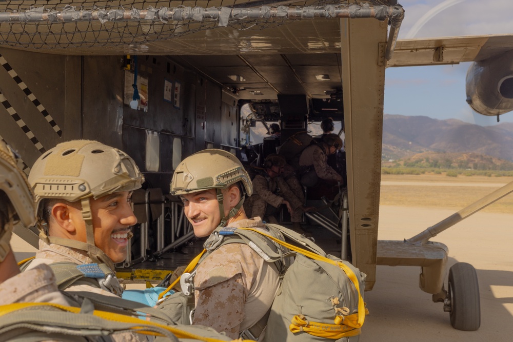 Two smiling military personnel in tactical gear and helmets are positioned at the rear ramp of a military transport aircraft, likely preparing for a parachute operation. Both are wearing parachute rigs with yellow harness components and MARPAT camouflage uniforms consistent with U.S. Marines. Additional personnel are visible seated inside the aircraft cabin, with a mountainous landscape visible in the background.