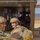 Two smiling military personnel in tactical gear and helmets are positioned at the rear ramp of a military transport aircraft, likely preparing for a parachute operation. Both are wearing parachute rigs with yellow harness components and MARPAT camouflage uniforms consistent with U.S. Marines. Additional personnel are visible seated inside the aircraft cabin, with a mountainous landscape visible in the background.