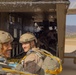 Two smiling military personnel in tactical gear and helmets are positioned at the rear ramp of a military transport aircraft, likely preparing for a parachute operation. Both are wearing parachute rigs with yellow harness components and MARPAT camouflage uniforms consistent with U.S. Marines. Additional personnel are visible seated inside the aircraft cabin, with a mountainous landscape visible in the background.