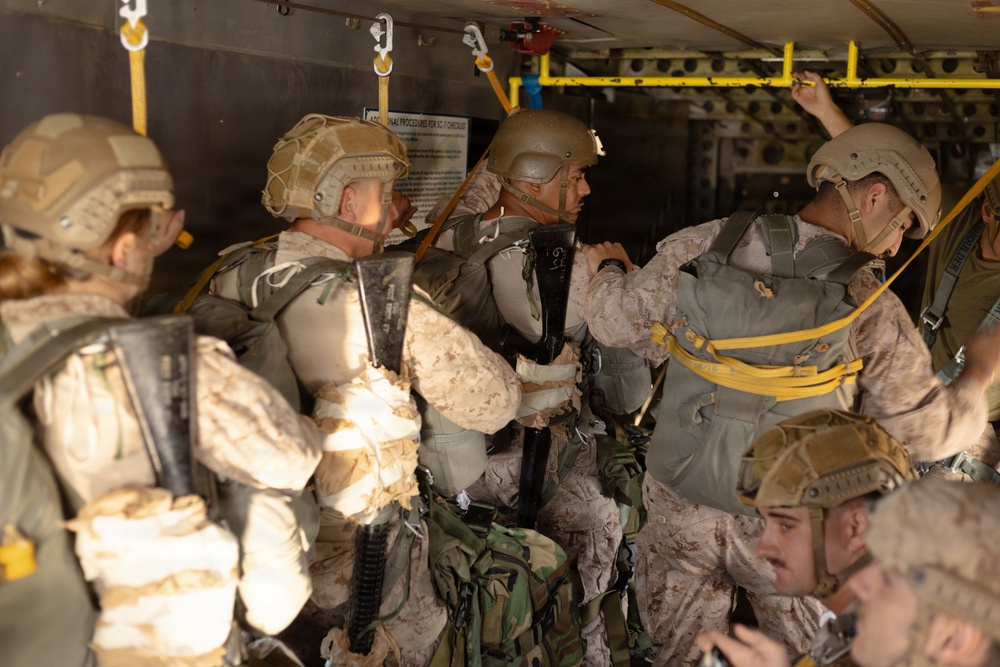 A group of military personnel in desert camouflage uniforms are inside what appears to be a military aircraft, equipped with parachute gear and combat helmets. The soldiers appear to be preparing for a parachute jump operation, with static lines and yellow anchor cables visible overhead. The scene depicts a pre-jump equipment check or staging sequence inside the aircraft cabin.