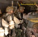 A group of military personnel in desert camouflage uniforms are inside what appears to be a military aircraft, equipped with parachute gear and combat helmets. The soldiers appear to be preparing for a parachute jump operation, with static lines and yellow anchor cables visible overhead. The scene depicts a pre-jump equipment check or staging sequence inside the aircraft cabin.