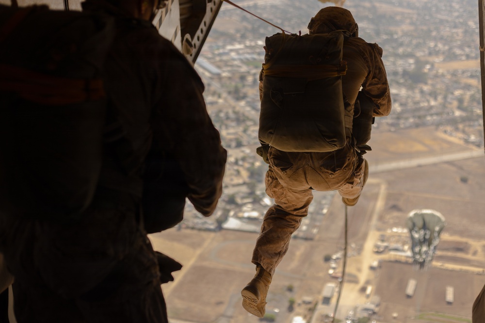 A military personnel in full combat gear is rappelling or fast-roping from what appears to be a helicopter at significant altitude over an urban/suburban landscape. A second individual is visible in the foreground, silhouetted against the open door or ramp of the aircraft. The terrain below shows a mix of agricultural fields, roads, and developed areas.