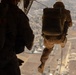 A military personnel in full combat gear is rappelling or fast-roping from what appears to be a helicopter at significant altitude over an urban/suburban landscape. A second individual is visible in the foreground, silhouetted against the open door or ramp of the aircraft. The terrain below shows a mix of agricultural fields, roads, and developed areas.