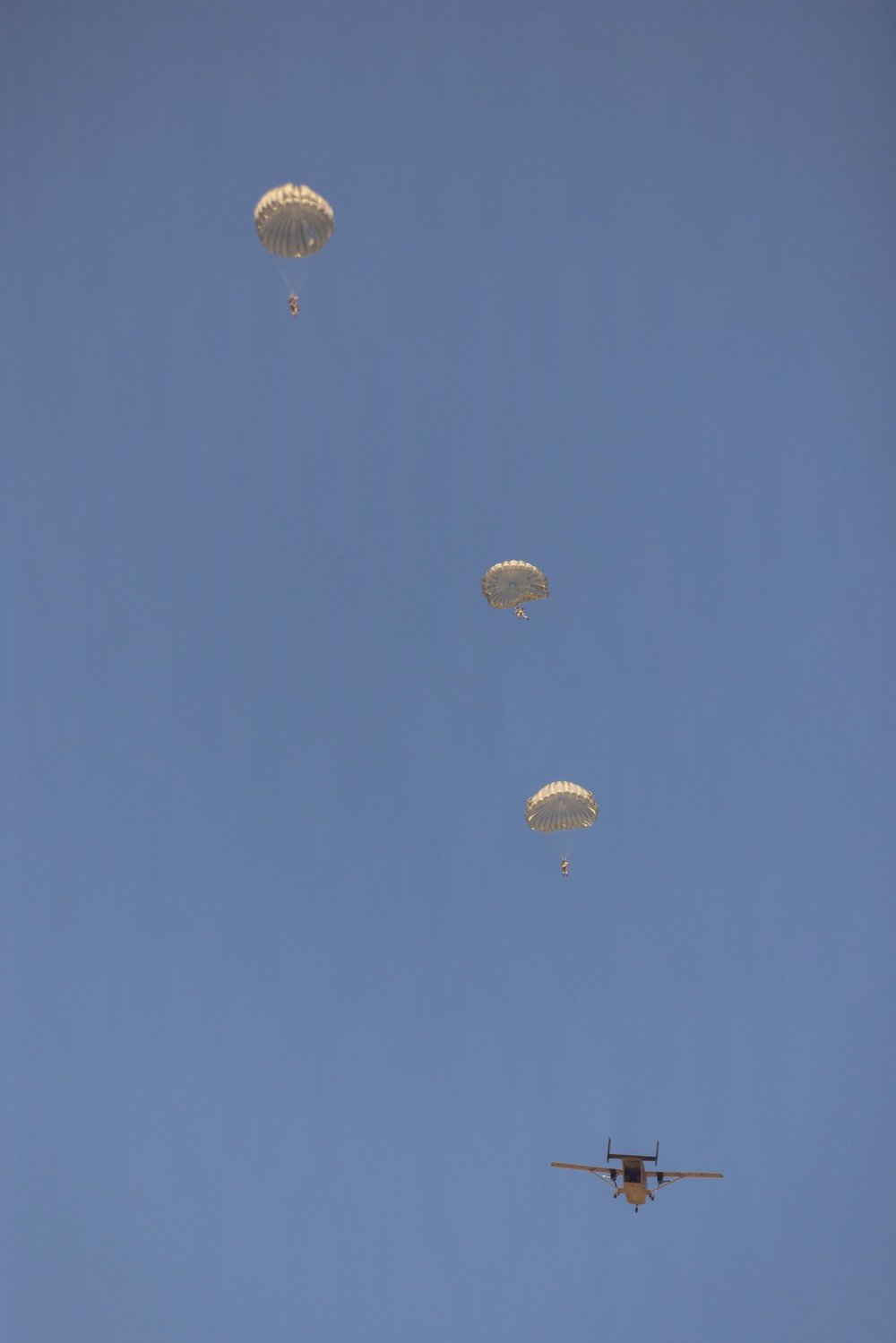 A military static-line parachute operation is captured against a clear blue sky, showing three fully deployed round parachutes at staggered altitudes with suspended jumpers beneath each canopy. A twin-engine turboprop aircraft, consistent with a CASA CN-235 or similar military transport, is visible in the lower right, having just completed its drop pass. The parachutes are tan/olive-colored, consistent with military-issue T-10 or similar round canopies.