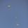 A military static-line parachute operation is captured against a clear blue sky, showing three fully deployed round parachutes at staggered altitudes with suspended jumpers beneath each canopy. A twin-engine turboprop aircraft, consistent with a CASA CN-235 or similar military transport, is visible in the lower right, having just completed its drop pass. The parachutes are tan/olive-colored, consistent with military-issue T-10 or similar round canopies.