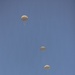 A military static-line parachute operation is captured against a clear blue sky, showing three fully deployed round parachutes at staggered altitudes with suspended jumpers beneath each canopy. A twin-engine turboprop aircraft, consistent with a CASA CN-235 or similar military transport, is visible in the lower right, having just completed its drop pass. The parachutes are tan/olive-colored, consistent with military-issue T-10 or similar round canopies.