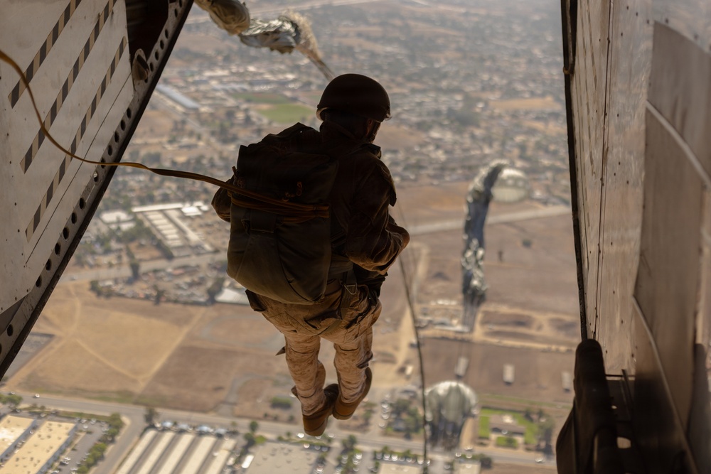 A military paratrooper is captured mid-jump from the open rear ramp of a large military aircraft, likely a CH-47 Chinook, at significant altitude over an urban and suburban landscape. The jumper is equipped with a static line parachute system, helmet, and full combat gear in desert camouflage. At least two other parachutes are visible already deployed in the background below.