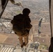 A military paratrooper is captured mid-jump from the open rear ramp of a large military aircraft, likely a CH-47 Chinook, at significant altitude over an urban and suburban landscape. The jumper is equipped with a static line parachute system, helmet, and full combat gear in desert camouflage. At least two other parachutes are visible already deployed in the background below.