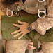 A close-up photograph of a person wearing desert digital camouflage (MARPAT pattern) military uniform and a parachute harness. The individual is holding or adjusting a green military equipment bag or parachute pack. A silver wristwatch is visible on the person's right wrist.