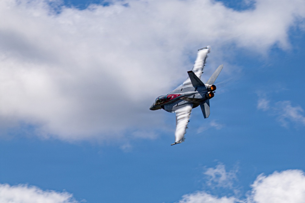 A military jet fighter is captured in a steep banking maneuver against a partly cloudy blue sky. The aircraft is generating prominent vapor condensation trails (vortices) streaming from its wingtips and fuselage, indicating high-G maneuvering at speed. The image appears to have been taken at an airshow or during a demonstration flight.