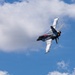 A military jet fighter is captured in a steep banking maneuver against a partly cloudy blue sky. The aircraft is generating prominent vapor condensation trails (vortices) streaming from its wingtips and fuselage, indicating high-G maneuvering at speed. The image appears to have been taken at an airshow or during a demonstration flight.