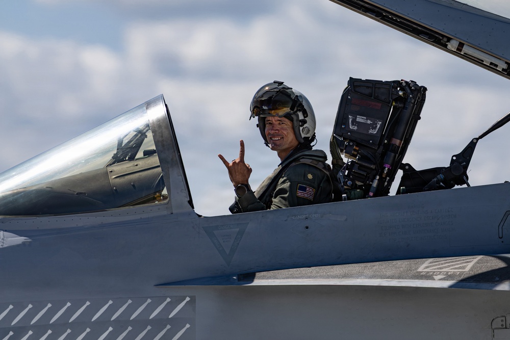 A military pilot is seated in the open cockpit of a fighter jet, smiling and making a hand gesture toward the camera. The pilot is wearing a flight suit with a visible US flag patch, a flight helmet, and appears to be on the ground at an airfield. The canopy is open and the ejection seat is visible behind the pilot.