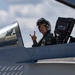 A military pilot is seated in the open cockpit of a fighter jet, smiling and making a hand gesture toward the camera. The pilot is wearing a flight suit with a visible US flag patch, a flight helmet, and appears to be on the ground at an airfield. The canopy is open and the ejection seat is visible behind the pilot.