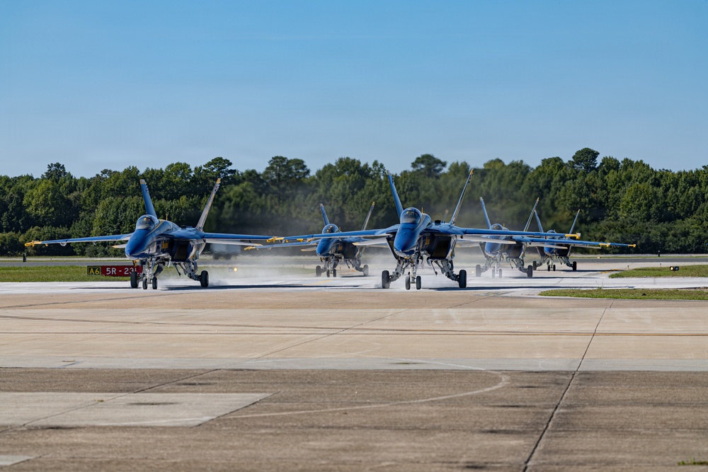A formation of Blue Angels F/A-18 Super Hornet jets taxi in close formation on an airfield runway. The aircraft are painted in the distinctive blue and yellow color scheme of the U.S. Navy Blue Angels flight demonstration squadron. Heat shimmer and jet exhaust are visible beneath the aircraft as they move along the tarmac.