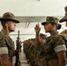 The image depicts a U.S. Marine Corps drill instructor wearing a campaign cover (drill sergeant hat) confronting or instructing a female recruit in MARPAT camouflage uniforms. The scene appears to take place in a covered outdoor or indoor training facility, with rows of recruits visible in the background. The interaction suggests an active training or inspection scenario typical of military basic training.