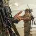 The image shows U.S. Marines in MARPAT camouflage uniforms during what appears to be a weapons handling or marksmanship instruction session indoors. A close-up foreground subject holds an M16/M4-style rifle while gesturing with their hand, suggesting they are explaining or demonstrating a technique. Additional Marines with rifles are visible in the blurred background, consistent with a training environment.