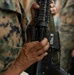 Close-up photograph of military personnel in woodland MARPAT (Marine Pattern) camouflage uniforms holding what appears to be an M16/M4-style rifle with a rail system and sling. A partial name tape reading 'SOSA' is visible on one uniform. The image focuses on the hands gripping the weapon, with multiple service members visible in the background.