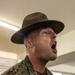 A U.S. Marine Corps Drill Instructor identified by the name tape 'NOVAK' is shouting with mouth wide open, wearing the distinctive campaign cover (drill instructor hat) with the Eagle, Globe, and Anchor emblem. He is dressed in MARPAT woodland digital camouflage utilities with a rank insignia visible on his collar. The image captures an intense moment consistent with drill instructor duties during recruit training.