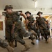 The image depicts what appears to be a U.S. Marine Corps drill instructor scenario, likely at a recruit training depot. Several personnel in MARPAT camouflage uniforms and campaign covers (drill instructor hats) are running and shouting in an indoor corridor. The motion blur on several figures suggests rapid movement and high-intensity activity consistent with recruit processing or training exercises.