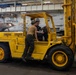 Two U.S. Navy personnel in hard hats are operating a large yellow industrial forklift inside what appears to be an aircraft carrier hangar bay. The forklift is marked with 'USN 13 19019' and has a Safe Working Load (SWL) of 20,000 lbs and a GVW of 31,000 lbs. Military aircraft, including jets marked with the number '214', are visible in the background.
