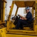A U.S. Navy sailor in blue coveralls and a hard hat operates a yellow shipboard forklift marked 'USN13 19019' inside what appears to be a ship's hangar or cargo bay. The forklift is rated for a safe working load of 20,000 lbs and is designated for shipboard use. The operator appears focused, looking to his right while seated at the controls.