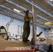 A military technician in olive drab uniform and green helmet/headgear stands atop an F-35B Lightning II aircraft (bureau number 305) inside what appears to be a naval vessel hangar bay. The technician is working on the vertical tail fin of the aircraft. A second F-35B (number 303) with a U.S. Air Force/Marine Corps roundel is visible in the background.