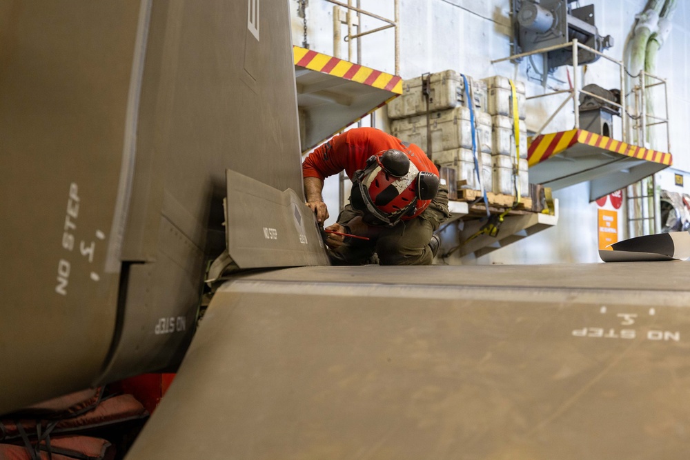 A technician wearing a red helmet and red shirt is performing maintenance or inspection work on a military aircraft, likely inside a hangar. The worker is crouched between large gray aircraft panels or fuselage sections marked with 'NO STEP' warnings. The environment appears to be a military aviation maintenance facility with yellow safety-marked platforms and equipment visible in the background.