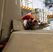 A technician wearing a red helmet and red shirt is performing maintenance or inspection work on a military aircraft, likely inside a hangar. The worker is crouched between large gray aircraft panels or fuselage sections marked with 'NO STEP' warnings. The environment appears to be a military aviation maintenance facility with yellow safety-marked platforms and equipment visible in the background.