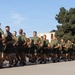 A large formation of military personnel in PT (physical training) uniforms are conducting a group run on a paved surface. One individual on the right side is wearing a blue shirt and carrying what appears to be a unit guidon or flag, suggesting a leadership or cadre role. The setting appears to be a military installation, likely a training base.