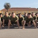 A large group of military personnel in olive green t-shirts and shorts are performing push-ups in unison on a paved surface. The setting appears to be a military base with a Spanish Colonial Revival-style building and palm trees visible in the background. The formation suggests a structured physical training exercise.