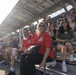 A crowd of spectators is seated in metal bleachers at what appears to be an outdoor sporting or public event. The stands are covered by a solar panel canopy roof, and attendees of various ages are watching something off-camera. The image is taken from a low angle near the front row, capturing both foreground subjects and a packed crowd behind them.
