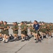A large formation of military personnel in olive drab PT (physical training) uniforms are conducting a group run on a parade deck or tarmac. One individual in a blue shirt, likely an instructor or drill sergeant, runs alongside the formation. The setting appears to be a U.S. military base, identifiable by the American flag on a flagpole and characteristic base architecture in the background.
