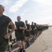A large formation of military personnel in physical training (PT) uniforms are running in a column on a wide paved surface, likely a parade deck or airstrip. They are wearing matching olive green t-shirts and shorts, consistent with U.S. Marine Corps recruit training attire. A single individual running alongside the formation on the right appears to be a drill instructor or pace-setter.