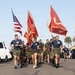 A group of U.S. Marines in physical training uniforms (navy blue shirts and olive green shorts) are running in formation through a parking lot while carrying flags. The flags visible include the U.S. national flag, U.S. Marine Corps colors, and what appears to be a '3rd Recruiting' battalion guidon. The setting appears to be a military installation, likely Marine Corps Recruit Depot (MCRD) San Diego, based on the architecture and palm trees.