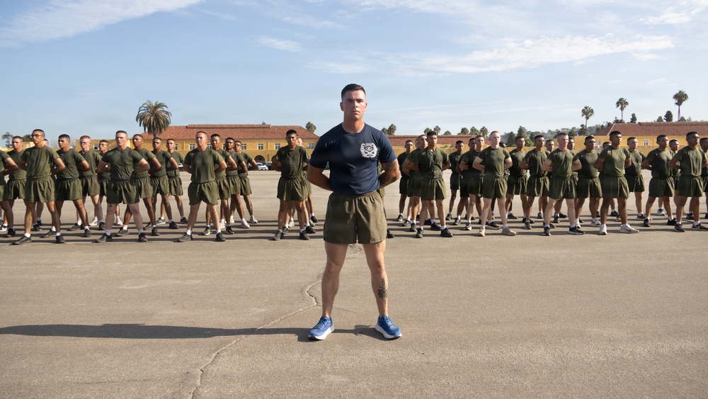 A military instructor or drill sergeant stands at the forefront of a large formation of recruits or trainees on a paved parade ground. The individuals in the background are uniformly dressed in olive green physical training attire and are standing in a disciplined formation with arms crossed. The setting appears to be a military base, likely Marine Corps Recruit Depot (MCRD) San Diego, based on the distinctive Spanish Colonial Revival architecture visible in the background.