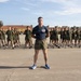 A military instructor or drill sergeant stands at the forefront of a large formation of recruits or trainees on a paved parade ground. The individuals in the background are uniformly dressed in olive green physical training attire and are standing in a disciplined formation with arms crossed. The setting appears to be a military base, likely Marine Corps Recruit Depot (MCRD) San Diego, based on the distinctive Spanish Colonial Revival architecture visible in the background.