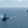 Pre-Commissioning Unit Ted Stevens, USS Daniel Inouye sail together Two naval warships are underway in open blue ocean waters under a partly cloudy sky. The foreground vessel is a U.S. Navy Arleigh Burke-class guided-missile destroyer, identifiable by the American flag and a blue pennant flying from its mast, with visible exhaust smoke trailing from its stack. A second warship of similar class is visible in the background to the right, both appearing to be on a parallel course.