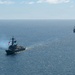 Two naval warships are underway in open blue ocean waters under a partly cloudy sky. The foreground vessel is a U.S. Navy Arleigh Burke-class guided-missile destroyer, identifiable by the American flag and a blue pennant flying from its mast, with visible exhaust smoke trailing from its stack. A second warship of similar class is visible in the background to the right, both appearing to be on a parallel course.